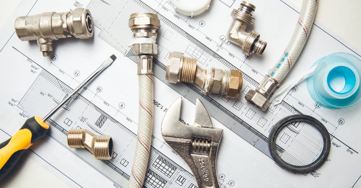 Two hoses, various fittings and connectors, a wrench, a screwdriver, and tape lie on top of construction blueprints.