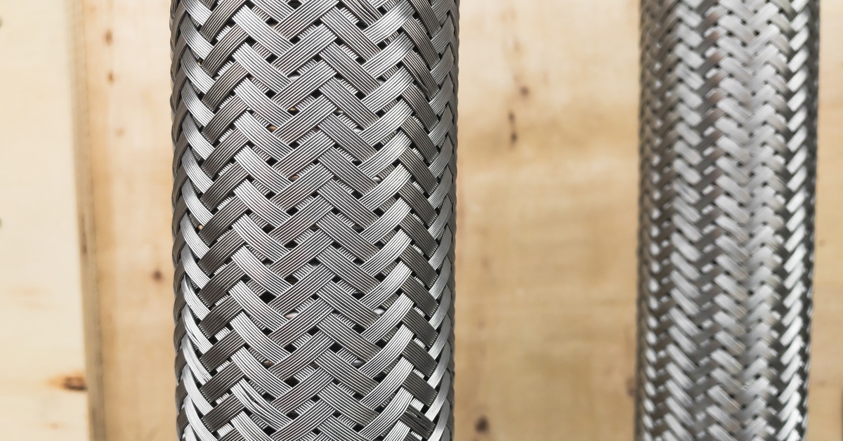 Two pressure hoses made of metal, with wood in the background. The hoses have a double braided stainless wire reinforcement.