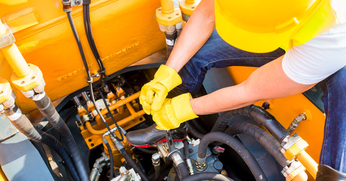 A construction professional wearing a yellow hard hat and gloves uses a tool to work on a construction vehicle.