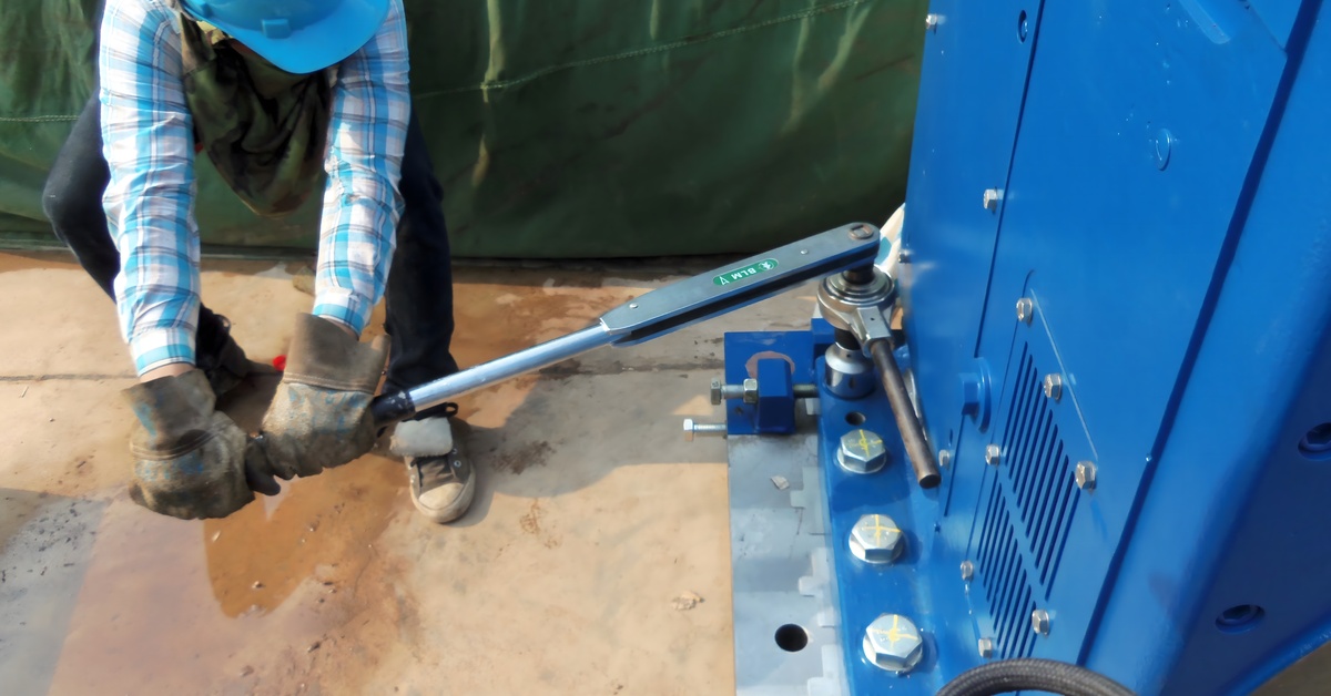 A person wearing a hard hat and work gloves uses a torque wrench to tighten parts on heavy machinery.