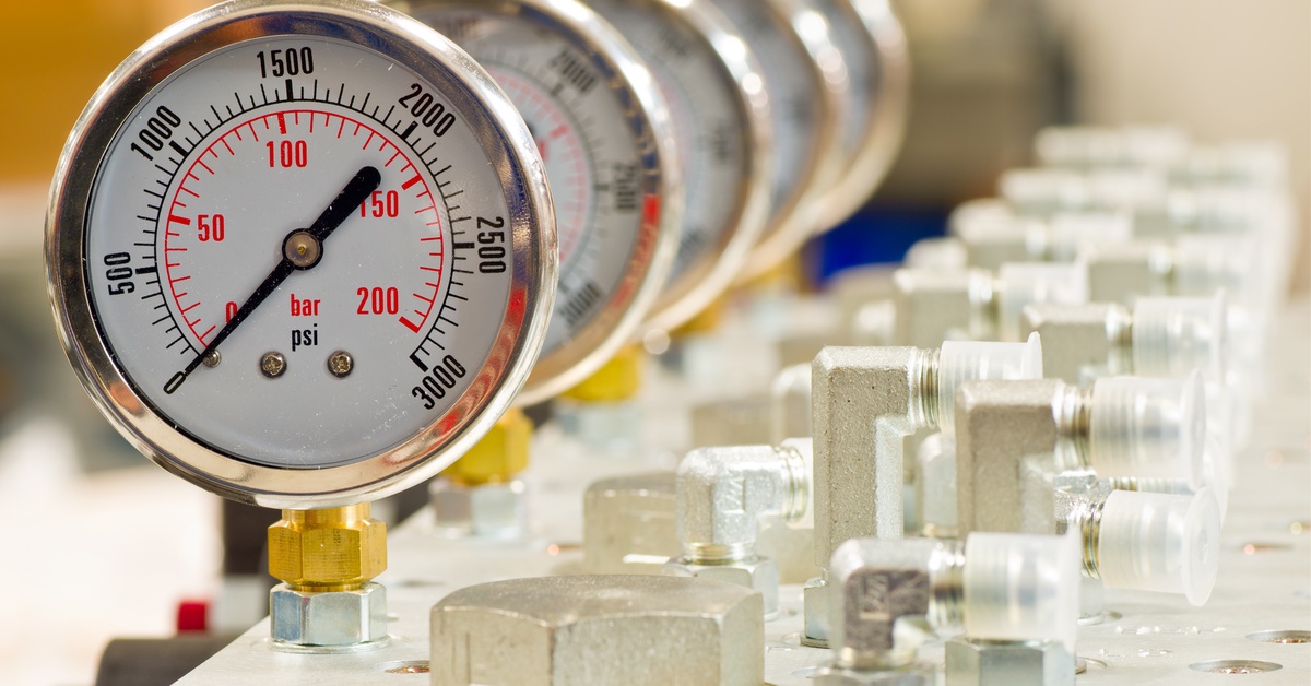 Five hydraulic pressure gauges lined up one behind the other on a table next to other hydraulic parts.