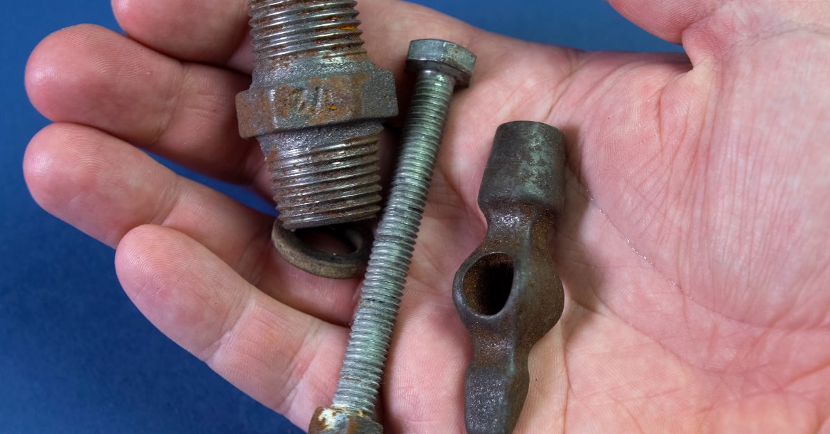A close-up of a person's right hand holding a small corroded hydraulic adapter and other metal items.