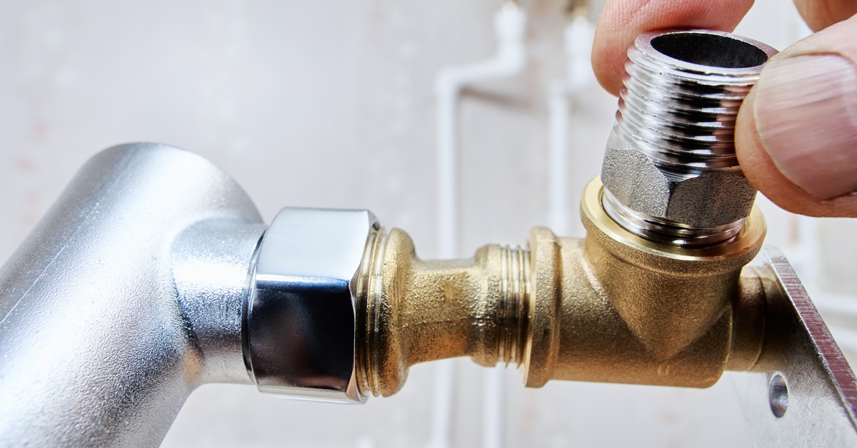 A person's thumb on a threaded reducer connected to a brass fitting in a plumbing system, with visible pipes in background.