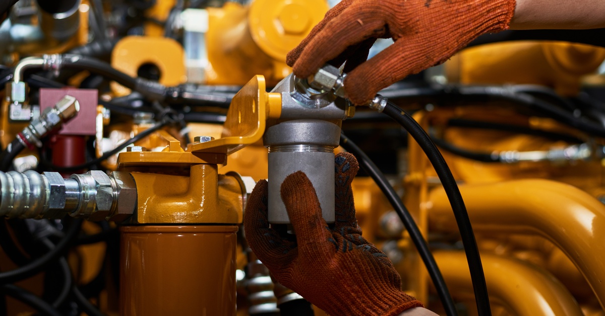 A close-up of a person wearing orange gloves holding components of an industrial hydraulic pump system.