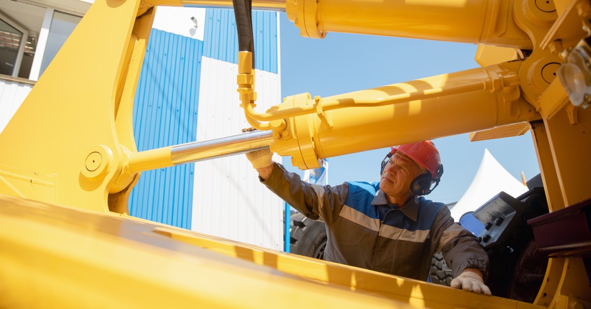 A male machinery tractor mechanic inspects the large yellow and silver hydraulic hose system equipment on the excavator.